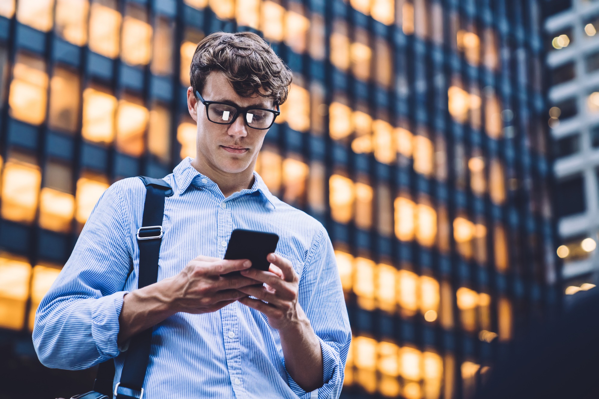 Man in shirt with rolled sleeves and glasses with bag texting on phone standing on background of lighted high building
